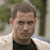 Pixelated image of a person with short brown hair wearing a black sleeveless top and light-colored outer garment, standing against a blurred green and gray background.