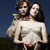 Two figures stand close together against a dark blue background; the figure in front wears a white off-shoulder top with visible straps and red accents.