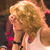 Pixelated close-up of a person with curly blonde hair wearing a light-colored sleeveless top, holding a dark object near their face.