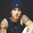 Pixelated image of a person in a dark sleeveless shirt and blue cap with visible tattoos, surrounded by pink pixelated text on a black background.