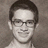 Static pixelated grayscale portrait of a person with short dark hair, glasses, and a neutral expression against a textured background.