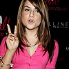 A person in a bright pink buttoned shirt making a peace sign with their right hand, wearing a pearl necklace and bracelets, against a dark background with partial text.
