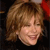 Pixelated close-up of a smiling person with light brown hair and a red accessory on the right side, set against a dark background.