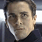Pixelated close-up of a person's face with short dark hair, light skin, and a dark collared shirt against a blurred background.