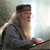 Pixelated image of a person with long hair and a beard wearing a dark cap and gray jacket, seated with a hand resting on a surface against a blurred background.