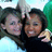 Two people with dark hair pose closely together, one holding a light-colored object near the other's face against a blurred background of green and blue tones.