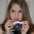 A person with long brown hair holds a black and silver digital camera up to their face, positioned in front of a light gray background.