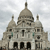 Pixelated image of a domed building with multiple spires, featuring a central dome, smaller domes, and dark rectangular windows against a light gray sky.