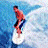 A person in red and black clothing rides a white surfboard on blue water, transitioning from standing to crouching with arms raised.