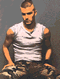 A person in a white sleeveless shirt and camouflage pants sits with hands on knees, head turned right to left