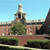 Pixelated static image of a building with a tall tower, red brick facade, green trees, and a clear blue sky background.