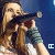A woman with long brown hair sings into a black microphone against a blue background, wearing a white top and green striped sleeves.
