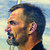 Pixelated profile view of a man with short dark hair, light skin, and a beard, facing left against a blurred light blue and yellow background.