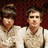 Two people in light-colored shirts sit side-by-side against a dark red background with yellow text reading "I <3 P!ATD" and "Take Me To The Hospital" on red and gray