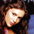 Pixelated close-up of a person with brown curly hair wearing a black top, showing partial face and shoulder against dark background with blue area.