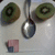 Two green circular objects with brown edges flank a silver spoon on a light gray background, with a small pink and blue shape below.