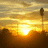 A pixelated sunset scene with a bright sun at the horizon, warm yellow and orange tones dominating the sky and water, and dark silhouettes of landmasses below.