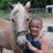 A pixelated static image shows a person with light hair wearing a dark blue shirt, standing next to a white horse in an outdoor setting with greenery and a vehicle in the background.