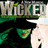 Pixelated image of a green-skinned character with dark hair and red lips, wearing a black hat and cloak, holding a white object against a black background.