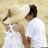 Two figures in white shirts and light-colored hats stand side-by-side against a beige background, with one figure's arm raised.