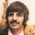 Pixelated image of a person with dark hair and mustache holding a light-colored object near their head.