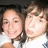 Two people in close proximity, one with dark hair and the other with lighter brown hair, both wearing white tops against a dark background.