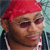 A pixelated close-up of a person wearing a red headwrap, dark sunglasses, and a dark jacket with light stripes, set against a blurred background.
