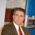Static pixelated image of a person in formal attire with a blurred background featuring blue and brown tones.