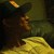 A person wearing a dark cap with a yellow logo sits on a couch with a yellow blanket, head turned slightly right in first frame, then left in second frame.