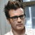 Pixelated close-up of a person with dark hair, wearing dark-framed glasses and a light-colored shirt, against a blurred background with vertical lines.