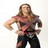 Pixelated static image of a person with long brown hair wearing a red and black patterned top and dark pants, standing against a plain white background.