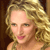 Pixelated close-up of a person with curly blonde hair, light skin, and pink lips against a dark brown background.
