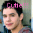 Pixelated image of a person with dark hair and light skin, wearing a blue garment, with pink text reading "Cutie!!!" above the head against a blurred background.