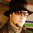 A person wearing a dark wide-brimmed hat, sunglasses, and a layered scarf, with a blurred background showing horizontal stripes.