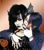 A static image of two figures with dark hair and pale skin, one holding a violin near the other's face against an orange background.