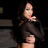 Pixelated image of a person with long dark hair wearing black clothing, holding an object near their mouth against a dark background.