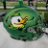 Pixelated green and brown creature with large white eye and yellow beak-like mouth against blurred green background.