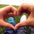 Two hands form a heart shape over a blurred outdoor background with green grass and blue clothing, surrounded by pixelated text reading "Oh," "It Is," and "Love."