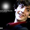 A close-up of a person with dark curly hair wearing a red-collared shirt, smiling broadly against a dark background with a bright light source on the left.