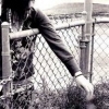 Black-and-white image of a person leaning against a chain-link fence, wearing a patterned dress and dark jacket, with a blurred background.