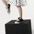 A woman with long blonde hair in a black top and silver checkered skirt performs dynamic poses against white background, including jumping on a black box.