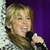 Pixelated image of a person with light brown hair holding a black microphone against a purple background with a white patterned edge.