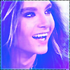 A close-up of a person with long light brown hair smiling broadly, showing teeth, under blue and purple lighting with a pixelated border.