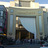 Pixelated static image of a building facade with columns and a large central opening, set against a light sky background.