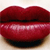 A close-up of glossy red lips with a subtle sheen, slightly parted to reveal a dark interior, set against a light beige background.