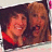 Two people in close proximity, one with dark hair and red clothing, the other with light brown hair, both smiling against a dark background.