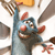 Blue cartoon rat with red nose and ears, smiling widely with teeth, holding a yellow object above its head, surrounded by brown and gray shapes.