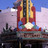 Pixelated image of a tall building with red and yellow vertical sections, blue sky background, and dark structures below.