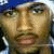 Pixelated close-up of a person's face with light brown skin tone, visible eyes and nose, mouth slightly open showing teeth, dark hair on top, blue background at upper left corner.