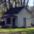 Pixelated image of a small house with dark roof and light walls, surrounded by trees and grass under a bright sky.